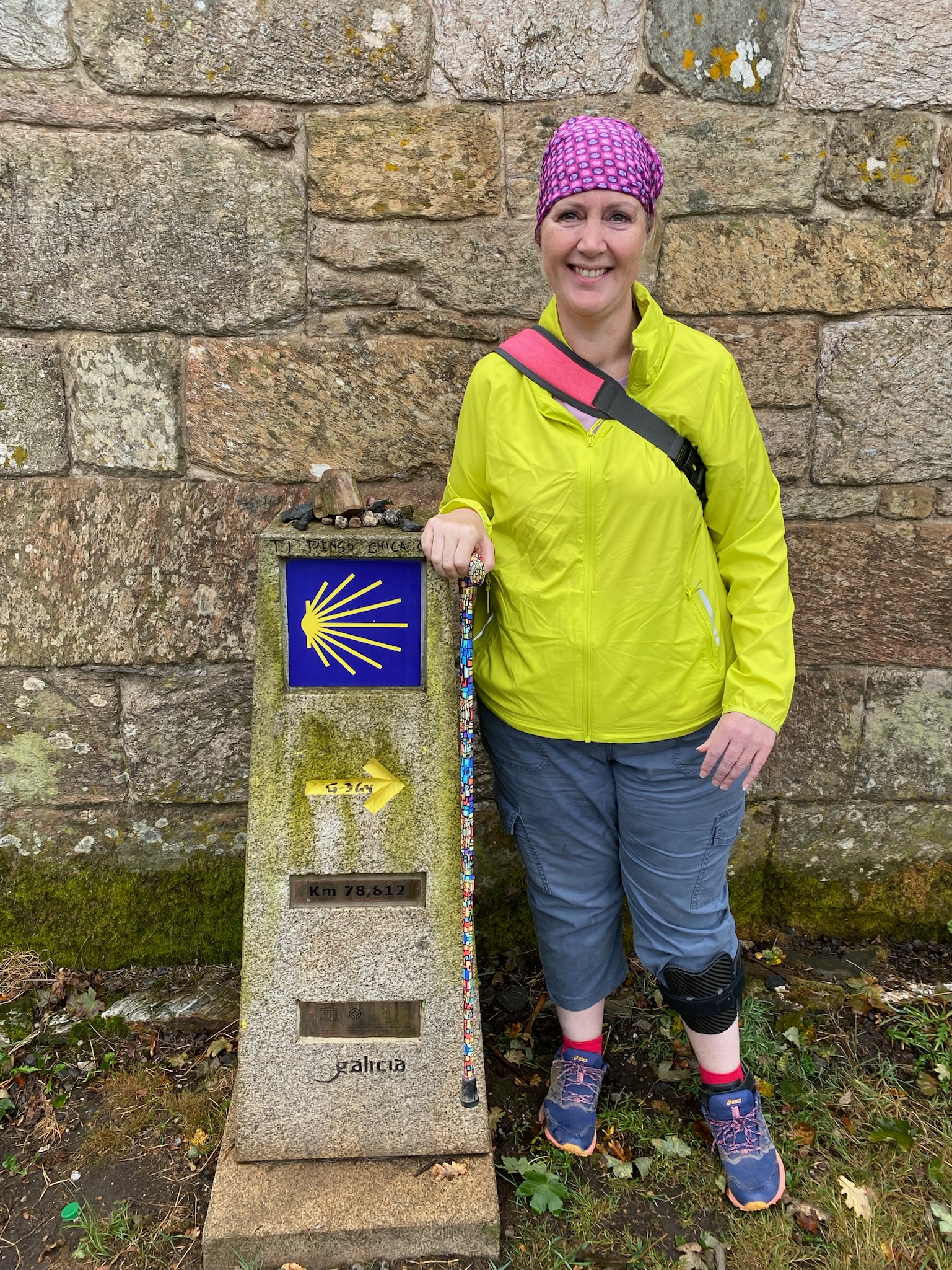 Sacha on the Camino in Galicia, leaning on a walking stick beside a stone marker with the yellow shell symbol, wearing an AFO ankle brace
