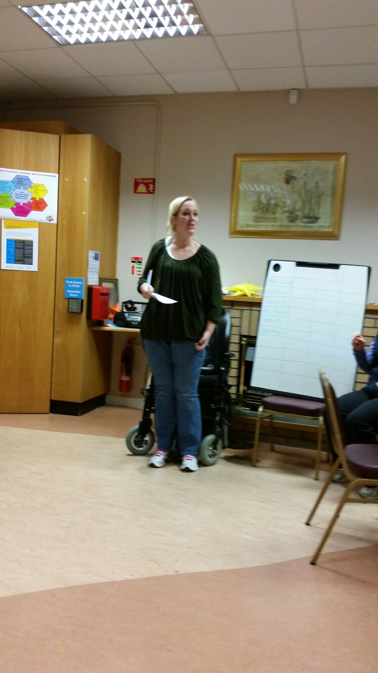 Sacha standing in front of her wheelchair, holding notes, in a rehabilitation room