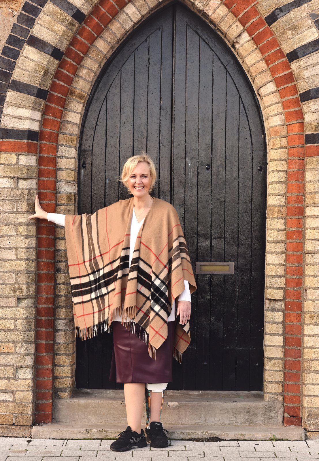 Sacha Dekker standing in front of a Gothic-arched black wooden door, wearing a brown striped wrap